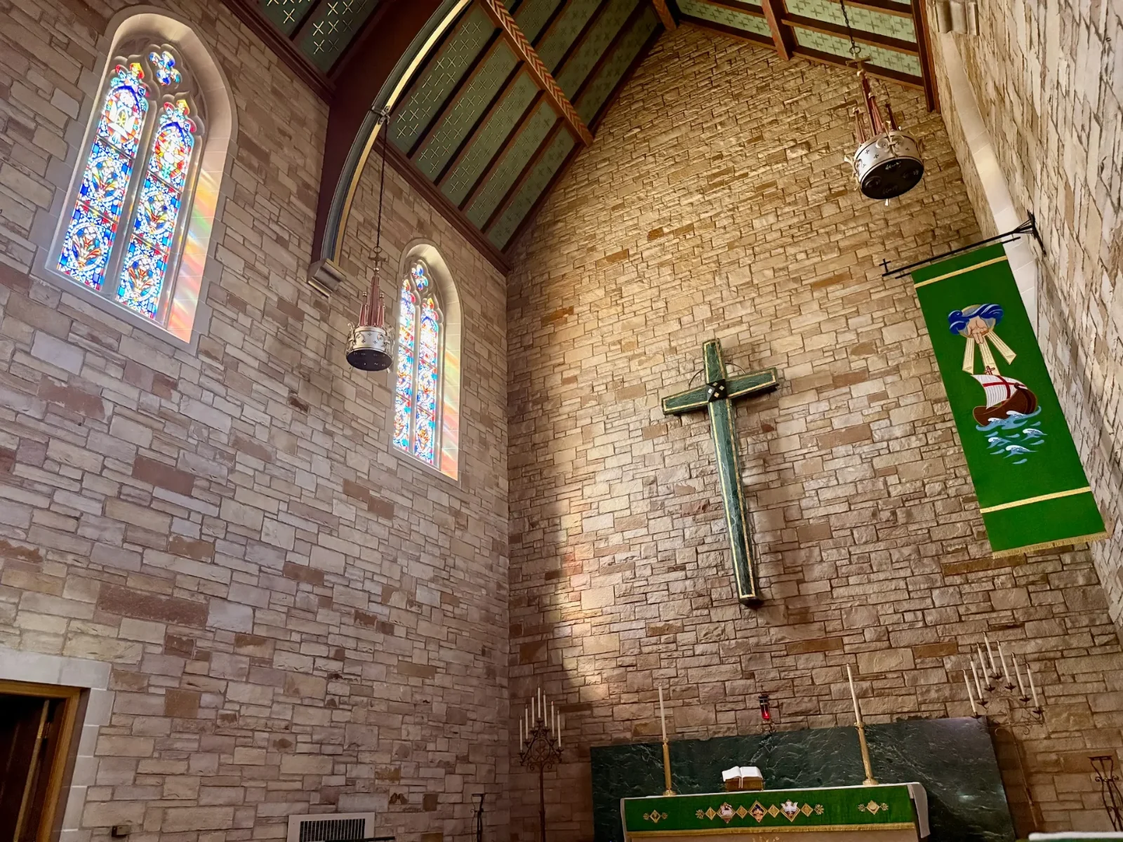 The chancel of Zion Lutheran Church in Wausau, with a green wooden cross on the stone wall beside stained-glass windows and the altar